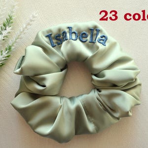 Puede incluir: Un scrunchie de satén verde claro con el nombre "Isabella" bordado en hilo azul. El scrunchie está disponible en 23 colores.