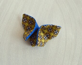 May include: A blue and yellow patterned bow tie. The bow tie has a blue backing and a yellow, blue, and brown floral pattern on the front. The bow tie is folded in a butterfly shape.