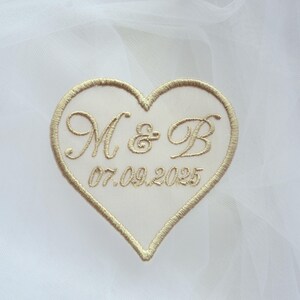 May include: A heart-shaped patch with gold embroidery. The patch features the initials "M & B" above the date "07.09.2025". The heart is outlined in gold thread and set against a white background.