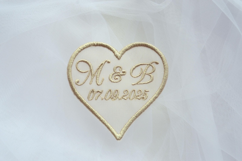 May include: A heart-shaped patch with gold embroidery. The patch features the initials "M & B" above the date "07.09.2025". The heart is outlined in gold thread and set against a white background.