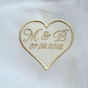 May include: Embroidered heart-shaped patch with gold thread. The patch features the initials "M & B" and the date "07.09.2025" in elegant script. The patch is set against a white, sheer fabric background, suggesting a wedding or special occasion.