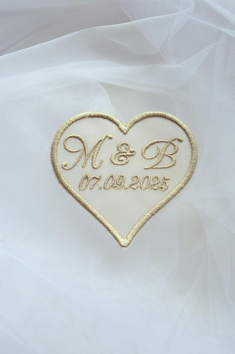 May include: Embroidered heart-shaped patch with gold thread. The patch features the initials "M & B" and the date "07.09.2025" in elegant script. The patch is set against a white, sheer fabric background, suggesting a wedding or special occasion.