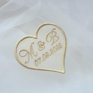 May include: A heart-shaped embroidered patch with the initials "M & B" and the date "07.09.2025" in gold thread. The patch is set against a white, sheer fabric background, suggesting a wedding or special occasion theme.