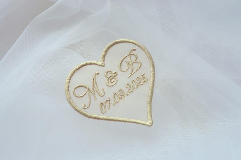 May include: A heart-shaped embroidered patch with the initials "M & B" and the date "07.09.2025" in gold thread. The patch is set against a white, sheer fabric background, suggesting a wedding or special occasion theme.