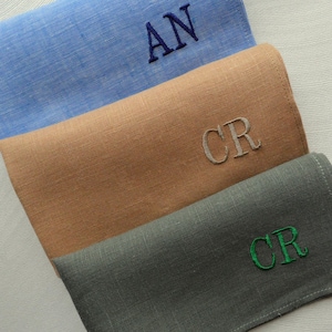May include: Three folded linen handkerchiefs in blue, tan, and grey, each embroidered with initials. The blue handkerchief features "AN" in navy thread, the tan one has "CR" in grey, and the grey one displays "CR" in green thread. The fabric has a subtle texture.