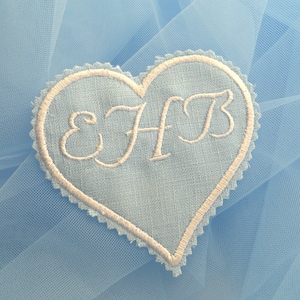 Puede incluir: Un parche de tela en forma de corazón azul claro con las iniciales bordadas en blanco "EHB" sobre un fondo de tul azul claro.