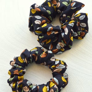 May include: Two black scrunchies with a leaf pattern in shades of brown, yellow, and blue. The scrunchies are made of fabric and are designed to hold hair in place.