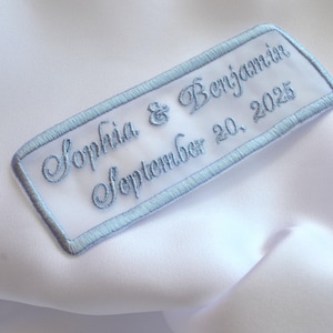 Könnte beinhalten: Weißer Stoff mit einem hellblau gestickten Aufnäher. Der Aufnäher zeigt die Namen "Sophia & Benjamin" und das Datum "20. September 2025" in eleganter Schrift, umrahmt von einem hellblauen Rand.