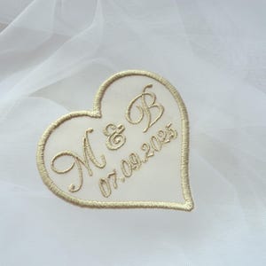 May include: A heart-shaped embroidered patch with gold thread on a white background. The patch features the initials "M & B" and the date "07.09.2025". The design is outlined with gold stitching.