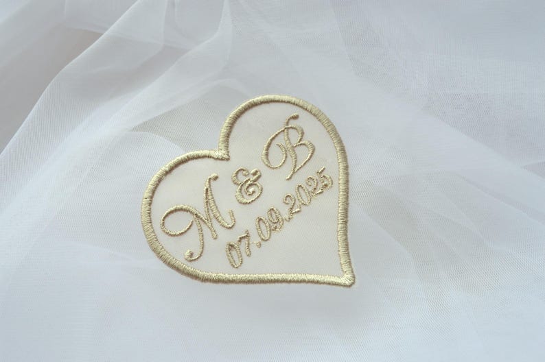 May include: A heart-shaped embroidered patch with gold thread on a white background. The patch features the initials "M & B" and the date "07.09.2025". The design is outlined with gold stitching.