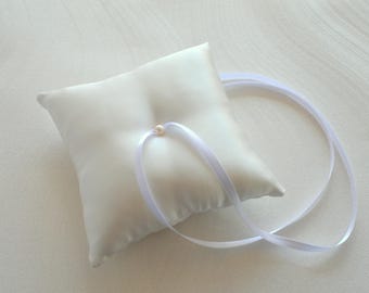 Könnte beinhalten: Ein weißes Satin-Ringkissen mit Perlenakzent und weißem Band. Das quadratische Kissen ist etwa 15 cm breit und dient zur Aufnahme von Eheringen. Das Band dient zur Sicherung der Ringe.
