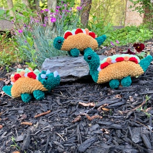 Puede incluir: Tres juguetes de dinosaurio de crochet con cuerpos verdes, espinas marrones y naranjas y ojos negros. Los dinosaurios están sentados en una cama de mantillo marrón.