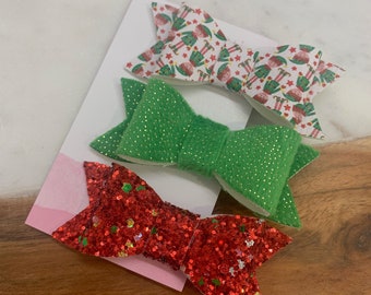 May include: Three Christmas-themed hair bows. One bow is red glitter, one is green, and one is white with a pattern of elves.