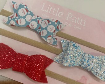 May include: Set of three Christmas-themed hair bows on nude nylon headbands. The bows are made of fabric with a Santa Claus print, red glitter fabric, and blue glitter fabric.