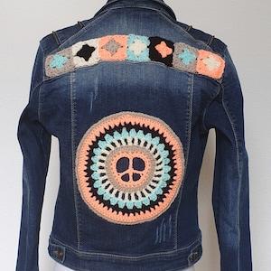 Op de afbeelding: Een blauwe denimjas met gehaakte patches op de rug. De patches zijn in tinten roze, perzik, beige, zwart en turquoise. De middelste patch is een grote cirkel met een vredesteken in het midden.