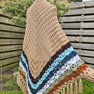 Puede incluir: Un chal tejido a crochet con una base beige y rayas de colores en marr&oacute;n, azul verdoso, blanco, verde y naranja. El chal tiene un borde de flecos.