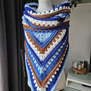 May include: A handmade, crocheted hooded cowl in shades of blue, white, and brown. The cowl features a geometric pattern with a triangular shape and a hood. The text "Annah Haakt Cozy Hooded Cowl" is visible on the side.