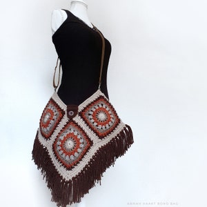 Op de afbeelding: Een gehaakte boho-tas met bruine franjes, met een geometrisch patroon van vierkanten met oranje en bruine accenten. De tas wordt gedragen over een bruin, mouwloos topje.