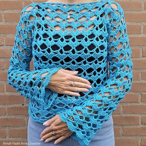 Op de afbeelding: Een turquoise gehaakte trui met lange mouwen en een boothals. Het open weefsel onthult de zwarte top die eronder wordt gedragen. De mouwen vallen wijd uit bij de polsen. De tekst "Annah Haakt Amira Sweater" is onderaan zichtbaar.
