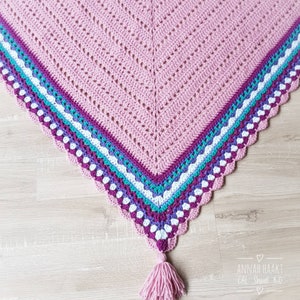 Op de afbeelding: Een handgemaakte gehaakte sjaal in een zachte roze tint, met een chevron ontwerp. De rand toont strepen in paars, turquoise en wit, met een geschulpte rand en een bijpassende roze kwast. Een gezellig accessoire.