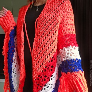 Puede incluir: Un chaleco poncho de ganchillo con un patr&oacute;n de rayas de colores en rojo, blanco y azul. El chaleco tiene un dobladillo con flecos y un cierre de corbata en el cuello. El chaleco est&aacute; hecho con un hilo ligero y tiene un ajuste holgado y fluido.