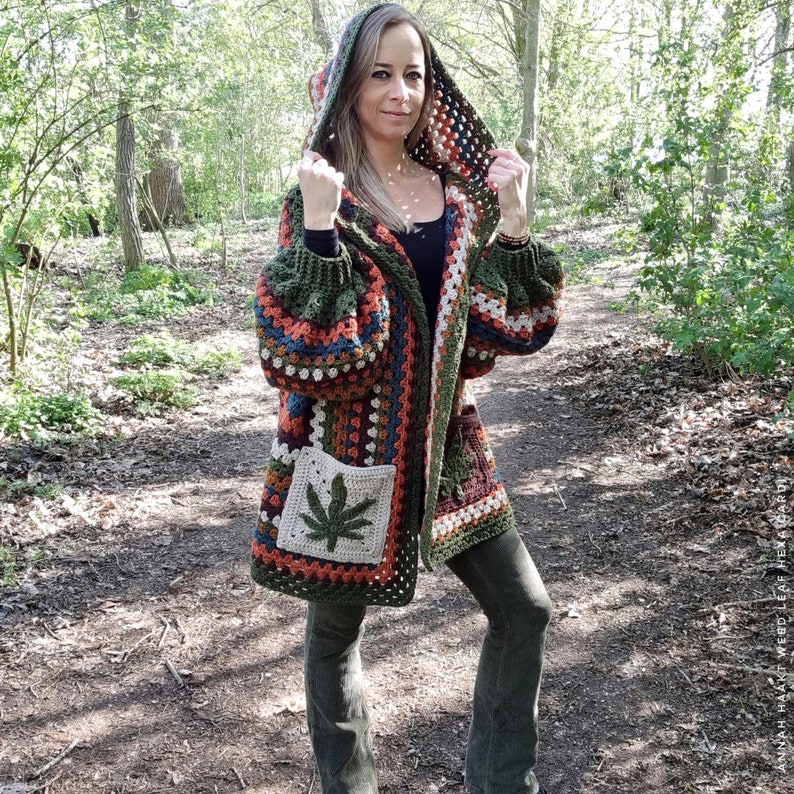 May include: A green, orange, and brown crocheted cardigan with a hood and a leaf design on the pocket. The cardigan is worn by a person standing in a forest.