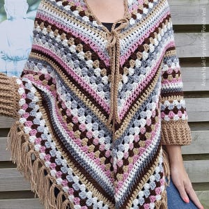 Op de afbeelding: Een gebreid poncho in een kleurenschema van bruin, roze, wit en grijs. De poncho heeft een franje rand en een strik sluiting aan de hals.