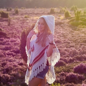 Op de afbeelding: Een crèmekleurige gehaakte poncho met capuchon, franjes en een rood-roze streepdessin. De poncho heeft een losse, open weefsel en wordt buiten gedragen. De achtergrond toont een veld met paarse bloemen.