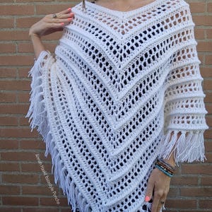 Op de afbeelding: Een witte gehaakte poncho met een V-hals en franjes. Het open weefpatroon zorgt voor een luchtige en stijlvolle uitstraling. De poncho is een veelzijdig kledingstuk, geschikt om te dragen als laagje of op zichzelf. De tekst "Annah Haakt Ibizastyle Poncho" is zichtbaar.