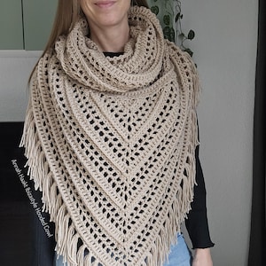 Puede incluir: Un chal con capucha de ganchillo beige con un borde con flecos. El chal tiene forma triangular y presenta un diseño calado. El texto "Annah Haakt Ibizastyle Hooded Cowl" es visible en el lado izquierdo de la imagen.