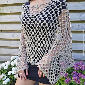Op de afbeelding: Een beige gehaakte poncho met klokmouwen. Het open weefselontwerp creëert een netachtig patroon. De poncho wordt over een zwarte top gedragen. De foto is buiten genomen met bloemen op de achtergrond.
