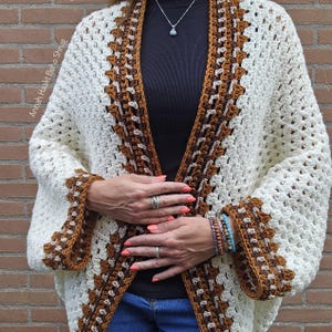 Op de afbeelding: Crèmegekleurde gehaakte boho-shrug met bruine rand. Het open vest heeft een geometrisch patroon en wijde mouwen. De tekst "Annah Haakt Boho Shrug" is zichtbaar aan de linkerkant van de afbeelding.