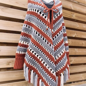 Op de afbeelding: Een gebreide poncho-vest met een gestreept patroon in de kleuren grijs, wit en oranje. De vest heeft een strik sluiting aan de hals en franjes aan de randen. "ANNAH HAAKT - HUGS & KISSES PONCHO VEST" staat onderaan de afbeelding.