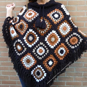 Op de afbeelding: Een zwarte gehaakte poncho met capuchon en franjes. De poncho heeft vierkante patronen in wit, bruin en oranje. De tekst "ANNAH HAAKT FALL VIBES PONCHO" is zichtbaar.