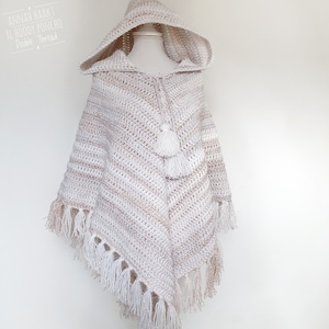 Op de afbeelding: Een gehaakt poncho met capuchon met een wit en beige gestreept patroon. De poncho heeft een kwast en franjes.