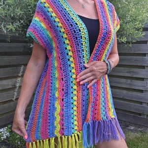 Op de afbeelding: Een mouwloze, aan de voorzijde open gehaakte kimono met verticale strepen in levendige regenboogkleuren zoals roze, geel, blauw en oranje. Het kledingstuk heeft een gestructureerd patroon en is afgewerkt met lange franjes aan de zoom.