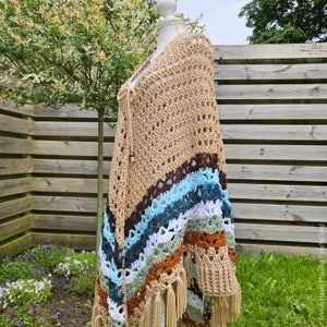 Puede incluir: Un chal tejido a crochet con una base beige y rayas de colores en tonos marrones, azules, verdes y blancos. El chal tiene un borde de flecos y se muestra en un maniqu&iacute;.