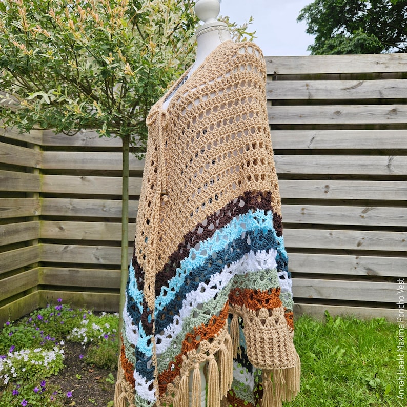 Puede incluir: Un chal tejido a crochet con una base beige y rayas de colores en tonos marrones, azules, verdes y blancos. El chal tiene un borde de flecos y se muestra en un maniqu&iacute;.