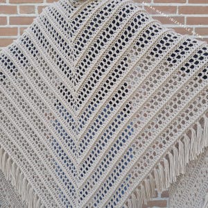 Op de afbeelding: Een beige gehaakte sjaal met een geometrisch openwerk ontwerp. De sjaal heeft een driehoekige vorm met franjes langs de onderkant. De tekst "ANNAH HAART IBIZASTYLE SHAWL" is rechtsboven zichtbaar.