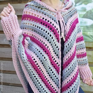 May include: A handmade crochet poncho in shades of pink, grey, and white. The poncho features a hood and long sleeves with a textured cuff. The design includes diagonal stripes and a tie closure with wooden beads.