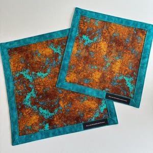 May include: Two square fabric mats featuring a mottled rust-colored and metallic gold pattern with vibrant turquoise accents. Each mat has a solid turquoise border and a small black rectangular label with the text HALLIESHANDMADEHAWKS.