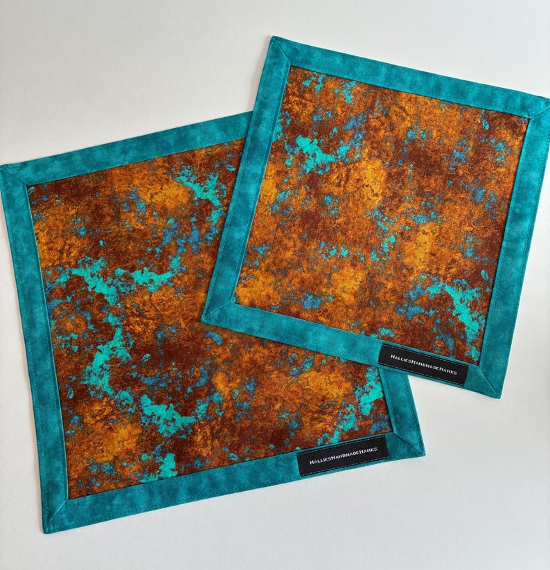 May include: Two square fabric mats featuring a mottled rust-colored and metallic gold pattern with vibrant turquoise accents. Each mat has a solid turquoise border and a small black rectangular label with the text HALLIESHANDMADEHAWKS.