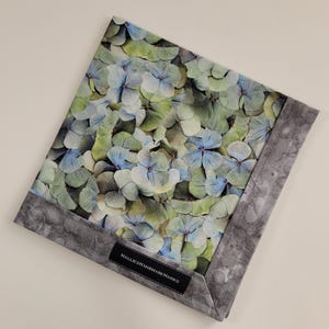 May include: A square fabric item with a floral pattern of blue, green, and white hydrangea blooms. The edges are bordered with a gray marbled fabric. The label reads "HALLIESHANDMADEHANKS".