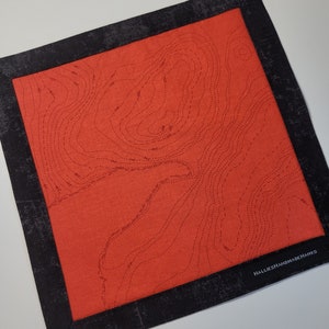 May include: A red fabric coaster with a black border and a topographic map design. The coaster has the text "HalliesHandmadeHanks" in the bottom right corner.