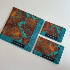 May include: Three square fabric cloths featuring a rust-colored and turquoise mottled pattern with a solid teal border. Each cloth has a black rectangular tag with the white text HALLIESHANDMADEHANKS sewn onto the bottom teal edge.