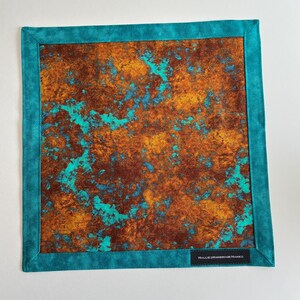 May include: A square fabric handkerchief featuring a central panel with a mottled, rust-colored and turquoise abstract pattern. The piece is framed by a solid teal border and includes a small black rectangular label reading HalliesHandmadeHanks.