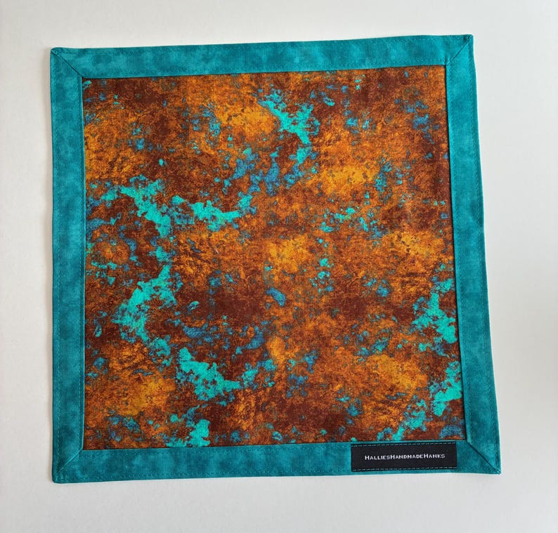 May include: A square fabric handkerchief featuring a central panel with a mottled, rust-colored and turquoise abstract pattern. The piece is framed by a solid teal border and includes a small black rectangular label reading HalliesHandmadeHanks.