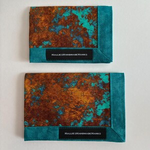 May include: Two rectangular fabric handkerchiefs featuring a mottled brown and turquoise pattern. Each piece has a solid turquoise border and a black rectangular label stitched to the front with the white text HALLIESHANDMADEHANKS.