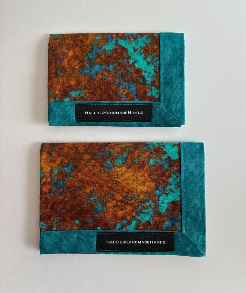 May include: Two rectangular fabric handkerchiefs featuring a mottled brown and turquoise pattern. Each piece has a solid turquoise border and a black rectangular label stitched to the front with the white text HALLIESHANDMADEHANKS.