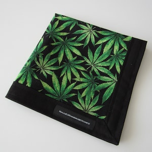 Könnte beinhalten: Eine schwarz-grüne Geldbörse mit einem Muster aus Cannabisblättern. Die Geldbörse hat einen schwarzen Rand und ein kleines Etikett mit dem Text "HALLIESHANDMADECHARES".
